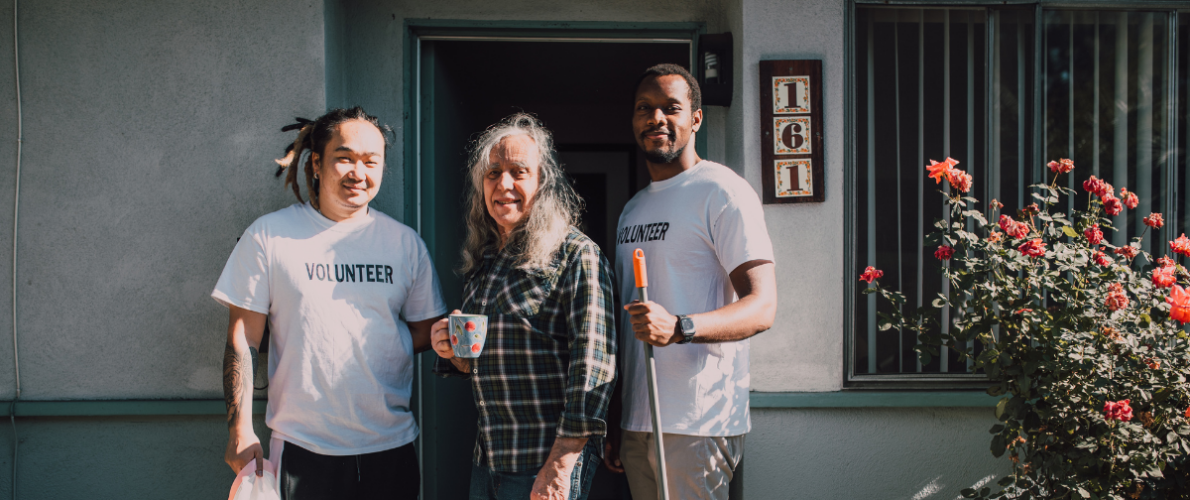 Two men wearing Volunteer t-shirts standing beside an older woman in front of a house.